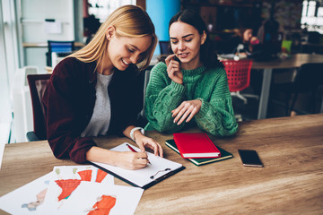 Fototapeta premium Cheerful two creative designers drawing sketch for new fashion project working at wooden desktop.Positive students collaborating on task sitting in university.Successful women teamworking in office