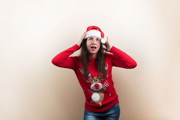 Young woman in Christmas knitted sweater with reindeer and Santa's hat holding head on beige background