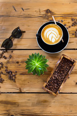 One cups of coffee on wood rustic background with beautiful latte art, green flower décor, roasted coffee beans   and sunglasses 