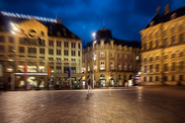 Fototapeta na wymiar Blurry motion image of man walking on Dam square in Amsterdam. Royal Palace is in the background. It is a rainy summer night with cloudy, dark blue color sky.