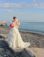 Fototapeta na wymiar Wedding by the sea. Beautiful bride with a red bouquet in a wedding dress at the water. Armenian bride on the stone beach in Batumi, Georgia. Concept marriage, just married