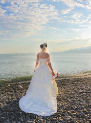 Fototapeta na wymiar Wedding by the sea. Beautiful bride with a red bouquet. Mountains and sea background. Bride looking at sunrise. Back view. Concept marriage, just married