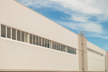 Facade of a long multi-span industrial building under a blue cloudy sky. Gray wall with staircase and tape glazing windows.