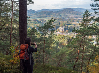 Young woman takeing cell phone picture of view on village Sloup v cechach with mediaval sand stone castle in luzicke hory, Lusatian Mountains with autumn colored tree forest and green hills, blue sky