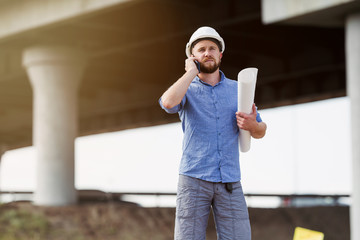 Brigadier in a white protective helmet talking on the phone with the architect on the background of the span of the bridge under construction