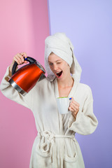 Winking woman pours water from kettle into cup. Girl pours water in mug from electric kettle. Happy housewife in bathrobe at home. Pretty girl pours hot water from kettle in kitchen. Girl making tea.