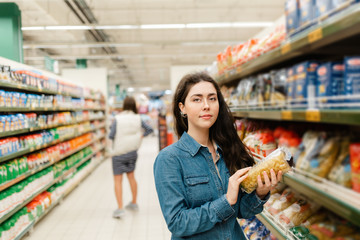 Shopping at the grocery store. Young beautiful woman in denim shirt holding a pack of pasta. In the background, the trading floor of the store