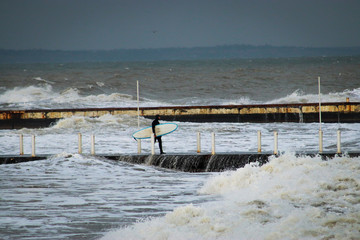 Surfer in a wetsuit with a surfboard on a city pier amidst powerful waves in Australia. Concept: ...