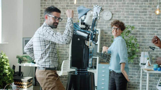 Joyful robotic engineers male and female are dancing with intelligent human-like robot in office enjoying business party. Work, artificial intelligence and fun concept.