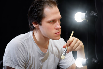 Obraz premium the actor sits in front of a mirror in a white t-shirt in a dark room and looks at his reflection, holding a brush for applying makeup to his face