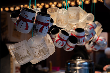 Cups of coffee. Christmas market in Europe. Transparent glass cups and white ceramic mugs in shape of snowmen hanging at dark background with blurry Christmas lights. Crockery utensil