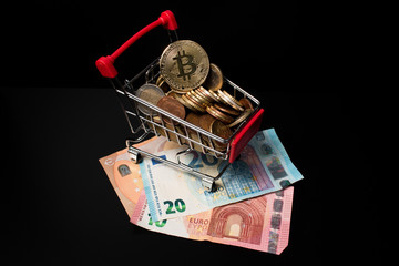 Miniature shopping cart filled with Euro coins and a bitcoin on the top placed on paper money with a black background. Making money with crypto currency.
