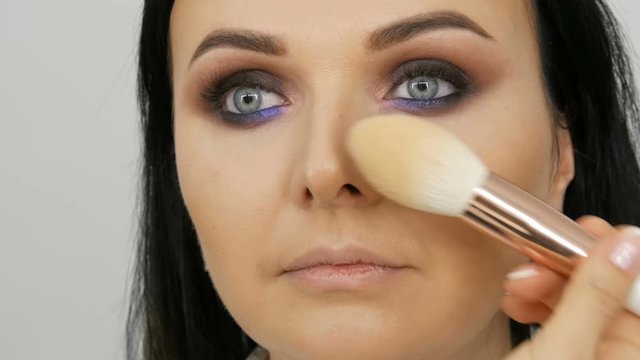 Makeup artist leads a woman in the face with a brush for applying powder, foundation or concealer. Lilac and pearly smoky eyes eyeshadow, eyes and face of woman close up. Professional high fashion.