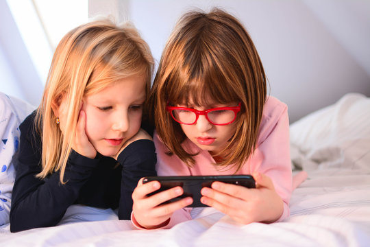 Front view of happy cute Caucasian little girls with mobile phone looking at cell phone on bed in a comfortable home. Two girls playing smartphones lying on a bed at mourning