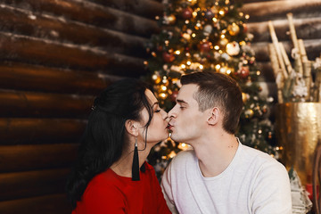 Beautiful young couple, boyfriend wearing sweater and girlfriend in sexy dress kissing in Christmas interior. Young woman in red dress and handsome man hugging and kissing in New year's interior