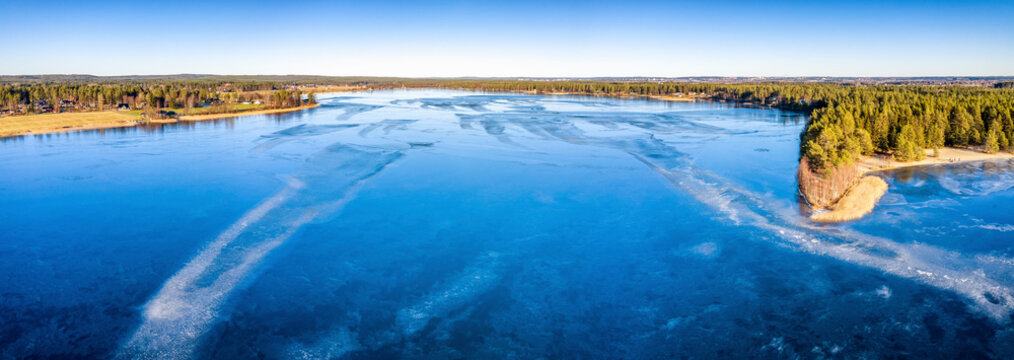 Aerial panoramic view over Swedish village and crystal clear frozen lake in Northern Scandianvia. Blue ice, clear skies, sunny winter day. Frozen lake like mirror, Umea city, Vasterbotten, Sweden