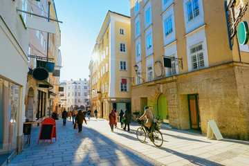 Fototapeta premium Kobieta na rowerze na ulicy starego miasta Salzburga w Austrii. Turyści i ludzie w mieście Mozart, Europa, zima. Panorama i punkt orientacyjny. Pejzaż miejski z rozbłyskiem słońca. Sklepy i sklep. Architektura budynku