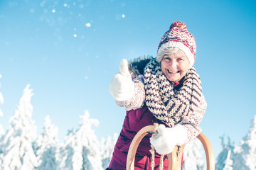 Mature woman with gray hair and winter cap is standing in beautiful winter landscape with sledge