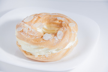Ring donut cut in the middle and inside whipped cream and on the top almond slices and sugar powder on white plate on bright background with space for text.