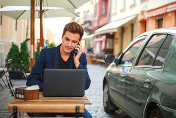 Young guy business man with laptop at table in summer cafe on street communicates on phone