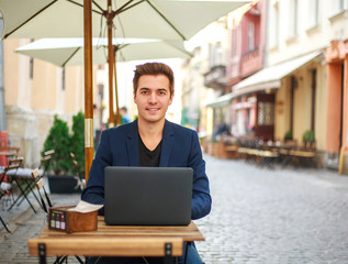 Young guy business man with laptop at table of summer cafe on street working outdoors