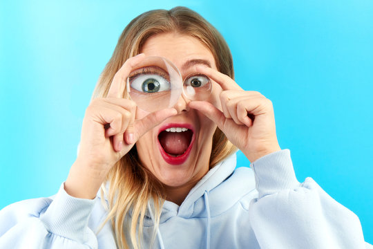 Fototapeta Young beautiful woman looking at the camera through magnifier with funny surprised expression, closeup isolated on blue. Enlarged eye, optical illusion, search