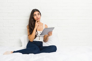 Young arab woman working with her laptop on the bed looking sideways with doubtful and skeptical expression.