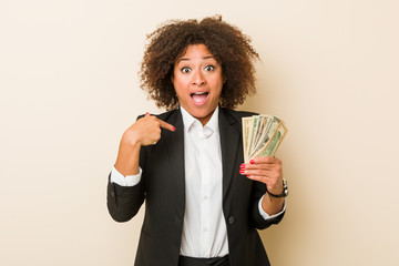 Young african american woman holding dollars surprised pointing at himself, smiling broadly.
