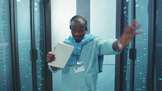 Ecstatic happy african engineer dancing in server corridor having fun flirting playing at camera with excitement. Smart handsome dancer in data center.