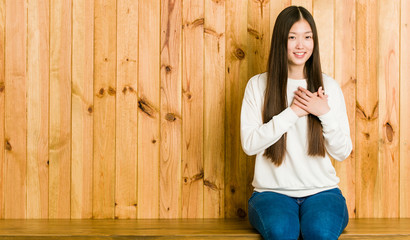 Young chinese woman sitting on a wooden place has friendly expression, pressing palm to chest. Love concept.