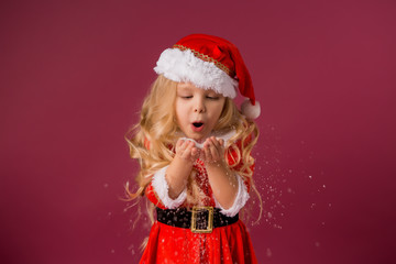 little blonde girl in a Santa suit blows snow off her hands. red background isolate. space for text