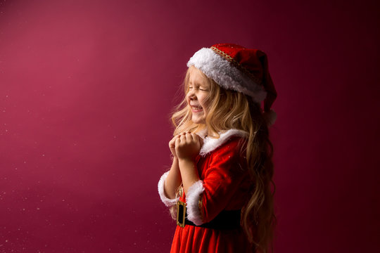 little blonde girl in a Santa costume holds her hands to her face and makes a wish.red background. space for text. Dreams come true. merry Christmas!