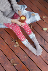 Woman with cup of tea sitting relaxing home in the window, she's covered in a blanket. Fall leaves on wooden patio deck. Autumn winter concept