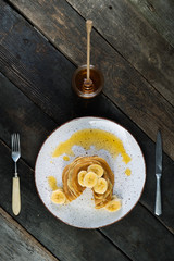 pancakes lie on a plate poured with honey with a honey spoon and decorated with bananas. plate with pancakes on an old wooden background.