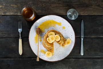 pancakes lie on a plate poured with honey with a honey spoon and decorated with bananas. plate with pancakes on an old wooden background.