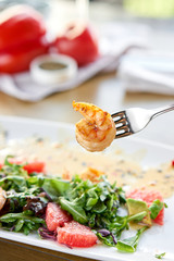 Close-up of grilled shrimps on a fork. Woman eats Delicious salad with slice grapefruit and arugula salad and spicy fried prawns. Restaurant menu, natural and organic food concept.