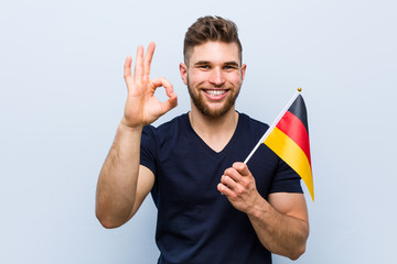 Young caucasian man holding a germany flag cheerful and confident showing ok gesture.