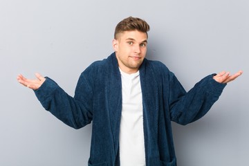 Young caucasian man wearing pajama doubting and shrugging shoulders in questioning gesture.