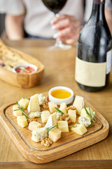 Wooden plate with cheese. Girl holding glass red wine on background . Delicious cheese mix with walnuts, honey. Tasting dish on a wooden plate. Food for wine.