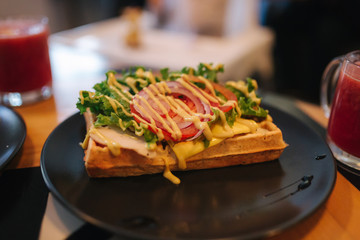 Waffles sandwich with bacon chiken and frech salad on black plate. Background of wooden table