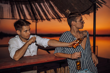 Two young friends drinking beer and pointing with hand to someone while enjoying in a good mood on the beach bar