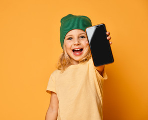 Smiling little girl kid in pink modern hat showing blank screen of mobile phone isolated over white background
