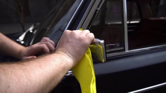 A car service worker polishes chrome parts on a blue old retro car with a yellow rag.