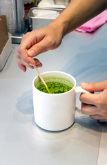Female barista hand stirring matcha green tea on a mug with a stick at a coffe-shop conter in Hiroshima.