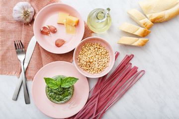 Raw pink fettuccine and Pesto sauce with ingredients: pine nuts, basil, olive oil, parmesan, garlic in pink plates with cutlery on white marble table.Making Spaghetti,pesto sauce. Top view, flat lay