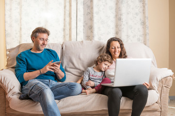 Child, a woman and a man sitting on a sofa watching a laptop and a smart phone.