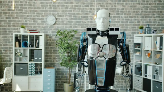 Portrait of smart robot rolling in office room looking at camera moving hand like waving hello. Modern technology, artificial intelligence and engineering concept.