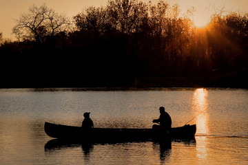Obraz na płótnie Canvas Close-Up Silhouette of a Man and a Child Together in a Canoe on a Lake in the Evening - with a Vibrant Orange Sunset in the Background