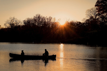 Fototapeta na wymiar Silhouette of a Man and a Child Together in a Canoe on a Lake in the Evening - with a Vibrant Orange Sunset in the Background