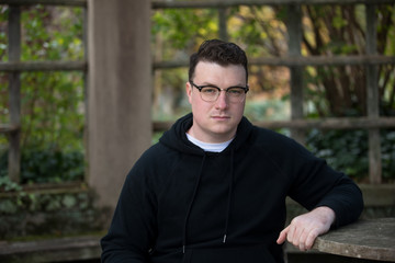 A caucasian male wearing a black hoodie sweater and glasses poses in a garden while surrounded by fall foliage on a cold afternoon.